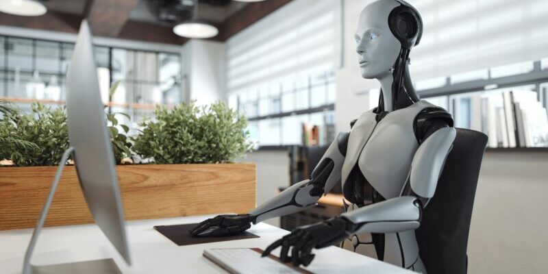 robot sitting at computer | ai trading models versus humans