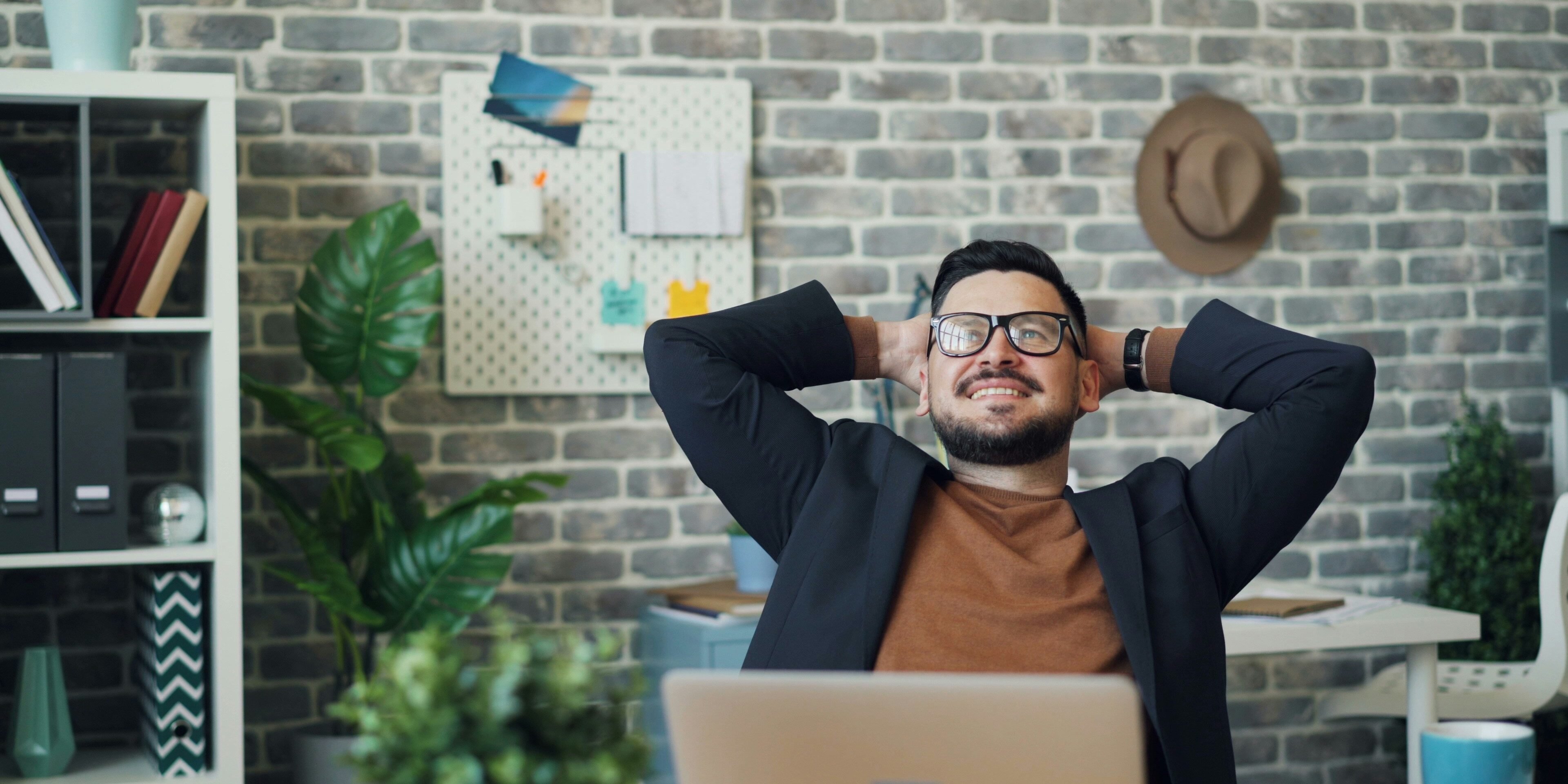 Man in glasses relaxing at his desk with hands behind head, smiling confidently—symbolizing complacency during periods of low market volatility.