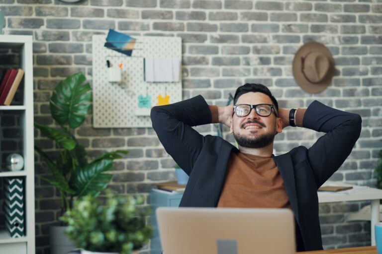 Man in glasses relaxing at his desk with hands behind head, smiling confidently—symbolizing complacency during periods of low market volatility.